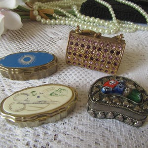May include: A selection of vintage trinket boxes in different shapes and designs. Includes a small, gold-toned purse-shaped box with purple gems, and oval boxes with blue, white, and decorated surfaces. Jewellery is in the background.