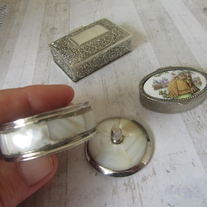 May include: An assortment of antique trinket boxes and a mother-of-pearl and silver ring box. The rectangular box has ornate silver detailing. The oval box features a painted scene. The ring box is open, revealing the interior.