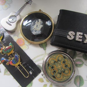 Può includere: Un assortimento di oggetti vintage tra cui un orologio a forma di chitarra bianca, un portafoglio nero con la scritta "SEXY" in strass, un cammeo rotondo, un compatto decorativo e un set di spille a forma di anatra colorata.