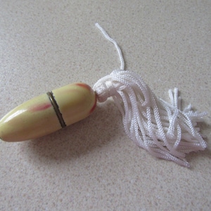 May include: A small, cream-coloured, egg-shaped thimble with a red and white stripe design and a gold metal band. It has a white tassel attached.