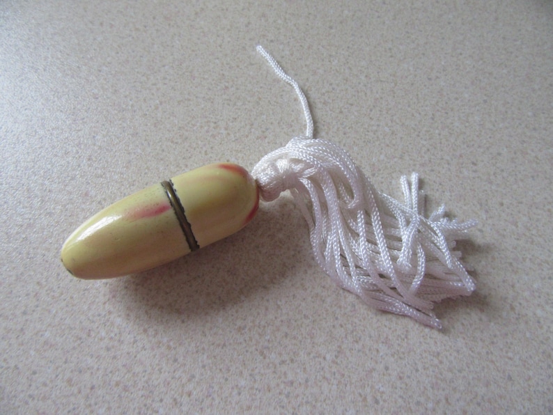 May include: A small, cream-coloured, egg-shaped thimble with a red and white stripe design and a gold metal band. It has a white tassel attached.