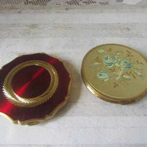 May include: Two vintage compacts. One is a red, scalloped-edge compact with a gold-coloured rope design around the centre. The other is a round, gold-toned compact with a floral design of blue roses.