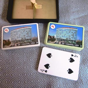 May include: A collection of vintage playing cards featuring a building with the text "MANCHESTER INNS" and "UK-CANADA-USA". The cards are stacked in two piles, with a single card showing the four of spades.