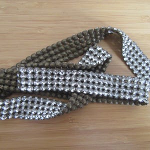 Op de afbeelding: Een zilveren en olijfgroene kralenriem of armband. Het zilveren gedeelte is gemaakt van kleine, sprankelende strass-steentjes. Het olijfgroene gedeelte is gemaakt van kleine, ronde kralen. Het item ligt op een lichtbruin houten oppervlak.