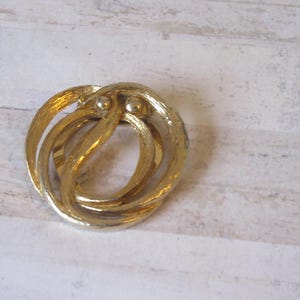 K&ouml;nnte beinhalten: Eine goldfarbene Brosche aus Metall mit einem wirbelnden, abstrakten Design. Die Brosche hat zwei kleine, runde Akzente.