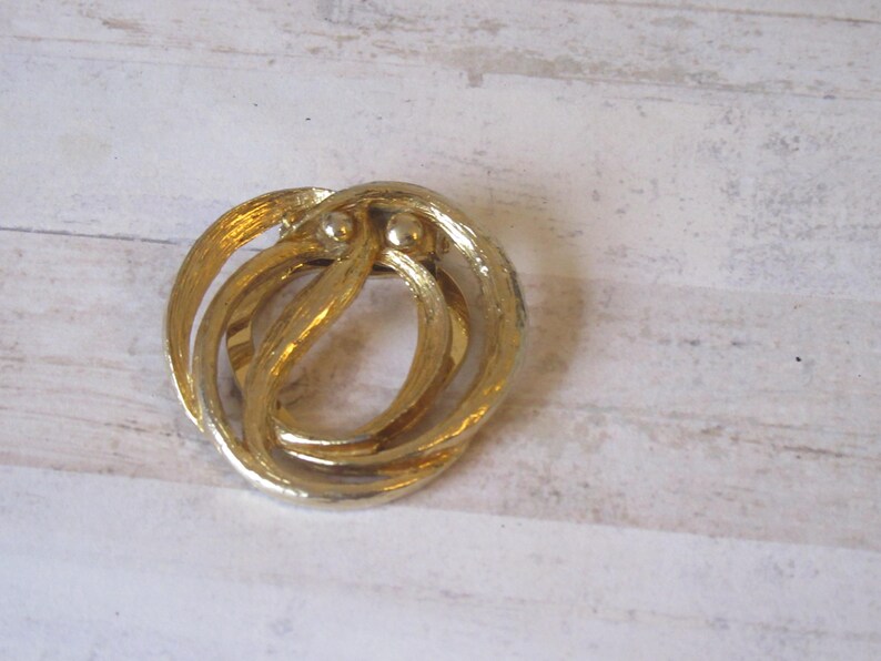 K&ouml;nnte beinhalten: Eine goldfarbene Brosche aus Metall mit einem wirbelnden, abstrakten Design. Die Brosche hat zwei kleine, runde Akzente.
