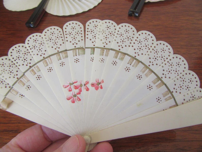 May include: A white folding fan with a delicate lace design and pink floral accents. The fan is made of paper and has a wooden handle.
