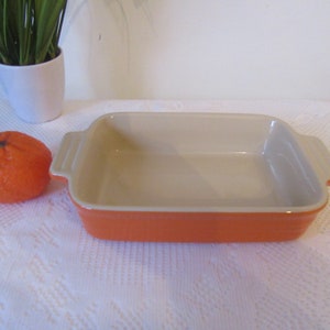 May include: A rectangular orange baking dish with a cream interior and two handles. It sits on a white surface. A small orange fruit is to the left, and a white pot with green plants is in the background. The dish is approximately 20 cm long.
