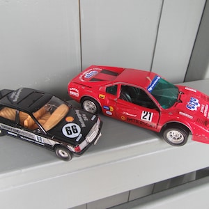 Könnte beinhalten: Zwei Spielzeug-Rennwagen. Einer ist schwarz mit goldenen Akzenten und der Nummer 66. Der andere ist rot mit der Nummer 21. Beide Autos haben Rennaufkleber und stehen auf einer grauen Oberfläche.