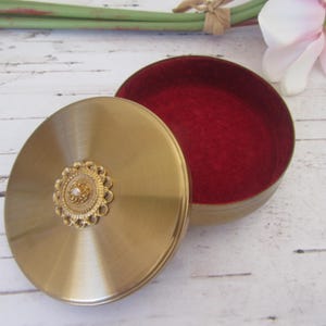 Peut inclure: Une boîte à bijoux ronde en métal doré avec un intérieur en velours rouge. Le couvercle est orné d'un motif floral décoratif avec un centre perlé.