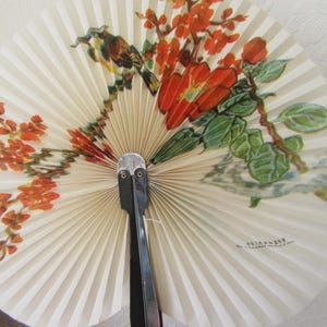 May include: A white folding fan with a black handle and a floral design featuring red flowers and green leaves. The fan has the text "K.F. KRAUSE" printed on it.