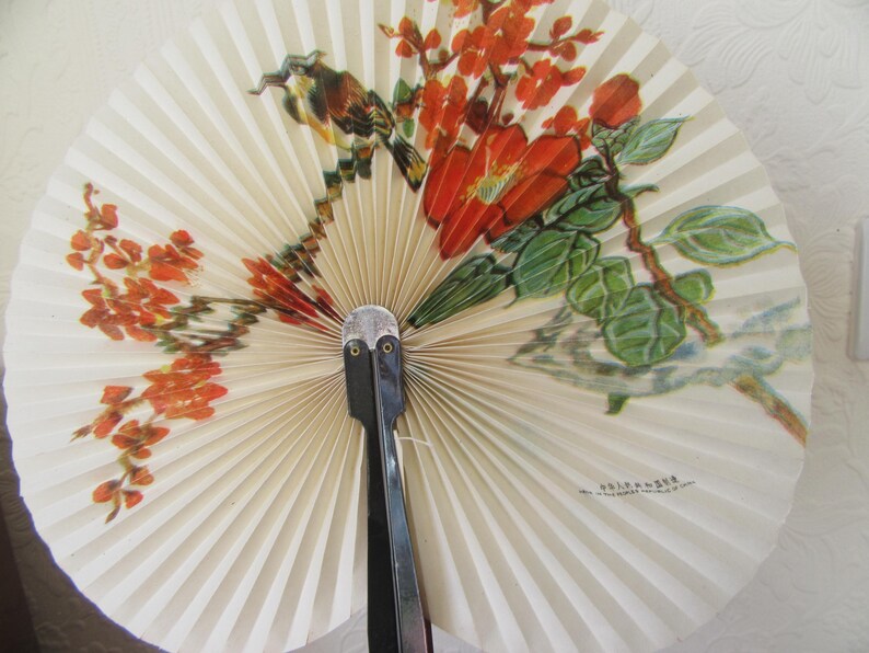 May include: A white folding fan with a black handle and a floral design featuring red flowers and green leaves. The fan has the text "K.F. KRAUSE" printed on it.