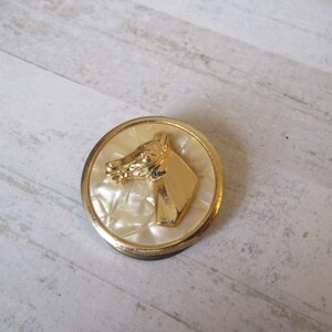 May include: A gold-toned button with a white, textured background featuring a raised image of a horse's head.