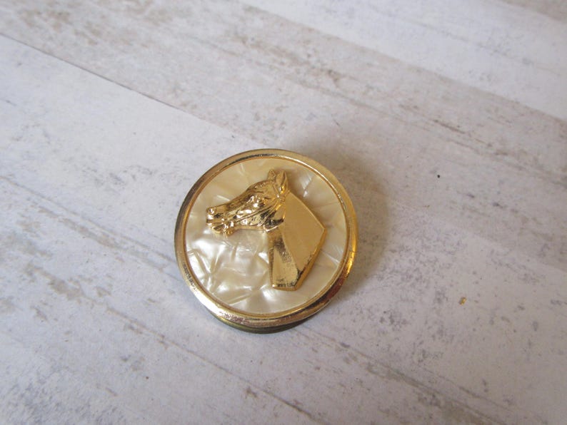 May include: A gold-toned button with a white, textured background featuring a raised image of a horse's head.
