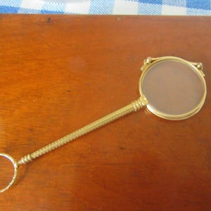 Op de afbeelding: Een goudkleurige vergrootglas met een ronde lens en een lange, getextureerde handgreep. De handgreep eindigt in een kleine ring. Het vergrootglas ligt op een houten oppervlak.