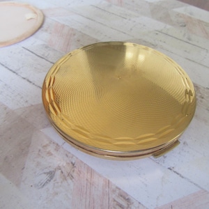 Op de afbeelding: Een rond, gouden compact met een getextureerd, swirlend ontwerp. Het compact heeft een scharnierend deksel en een klein slot.