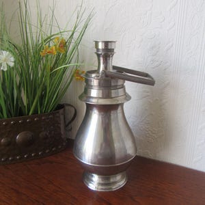 Op de afbeelding: Een zilveren metalen fles met een handvat en een tuit. De fles heeft een rond lichaam en een lange, smalle hals. De fles staat op een houten oppervlak.