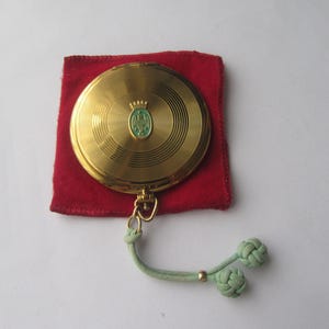 Könnte beinhalten: Ein goldener Kompakt-Spiegel mit einem grünen und goldenen Wappen auf dem Deckel. Der Spiegel befindet sich in einem roten Samtbeutel und hat einen grünen Lederquaste.