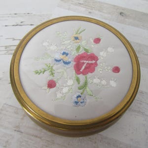 May include: A round, gold-colored metal trinket box with a white satin interior. The lid is decorated with a floral embroidery design in shades of pink, blue, green, and white.
