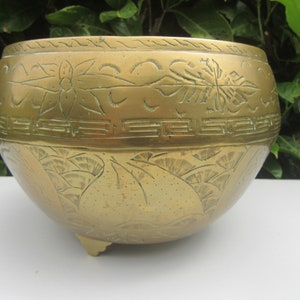 May include: A brass bowl with intricate engraved designs. The bowl has a wide rim and a rounded bottom with three small feet. The design features a repeating pattern of stylized flowers and leaves.