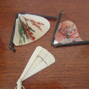 May include: Three decorative folding fans with intricate designs. One fan is white with a floral pattern, another is pink with a floral pattern, and the third is white with a geometric pattern.