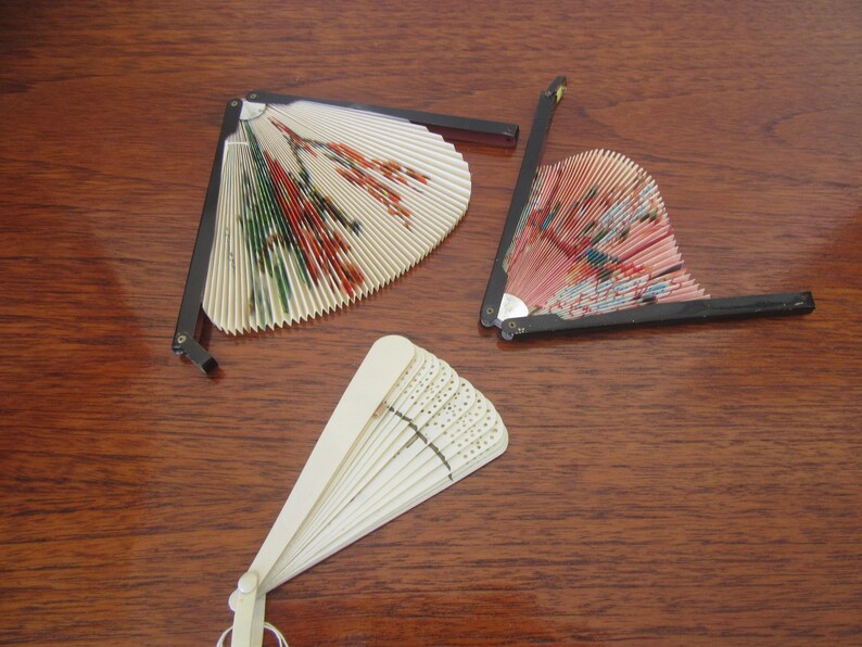 May include: Three decorative folding fans with intricate designs. One fan is white with a floral pattern, another is pink with a floral pattern, and the third is white with a geometric pattern.