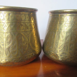 May include: Two antique brass pots with intricate engraved designs. The pots are round with a wide base and a narrow opening. The designs are in a traditional Arabic style.