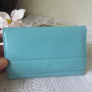 May include: A light blue leather wallet with a rectangular shape. The wallet has a flap closure and is accented with stitching. A white flower and a string of pearls are in the background.