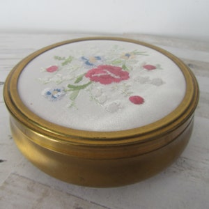 May include: A round, gold-colored metal trinket box with a white satin lid featuring an embroidered floral design in pink, blue, and green.
