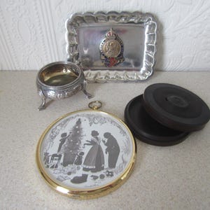 May include: A collection of vintage silver and gold-toned decorative items. A small silver bowl with three legs, a silver tray with a decorative crest, a round gold-toned frame with a silhouette image of a family around a Christmas tree, and a black wooden box.