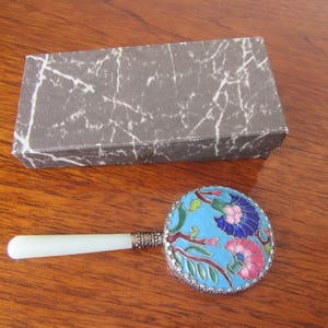 May include: A vintage magnifying glass with a round, floral-patterned head in shades of blue, pink, and green, and a white handle. A rectangular box with a marble-like pattern is also present. The items are on a wooden surface.