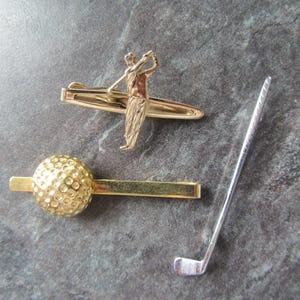 May include: Two gold-toned tie clips and one silver-toned golf club pin. One tie clip features a three-dimensional golfer in a swing pose, while the other tie clip is topped with a textured, spherical golf ball design. The golf club pin is slender.