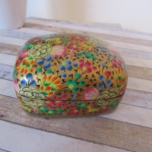 Könnte beinhalten: Eine kleine, quadratische, dekorative Schachtel mit goldenem Hintergrund und einem bunten Blumenmuster. Die Schachtel ist mit Blumen in den Farben Rosa, Gelb, Blau, Grün und Orange bemalt.