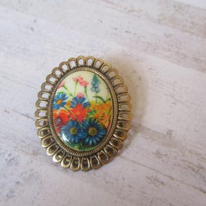 May include: A gold-toned oval brooch with a floral design. The brooch features a white background with colorful flowers in shades of blue, red, yellow, and pink.