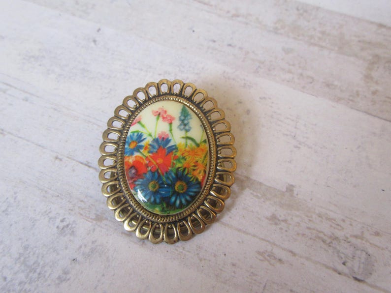 May include: A gold-toned oval brooch with a floral design. The brooch features a white background with colorful flowers in shades of blue, red, yellow, and pink.