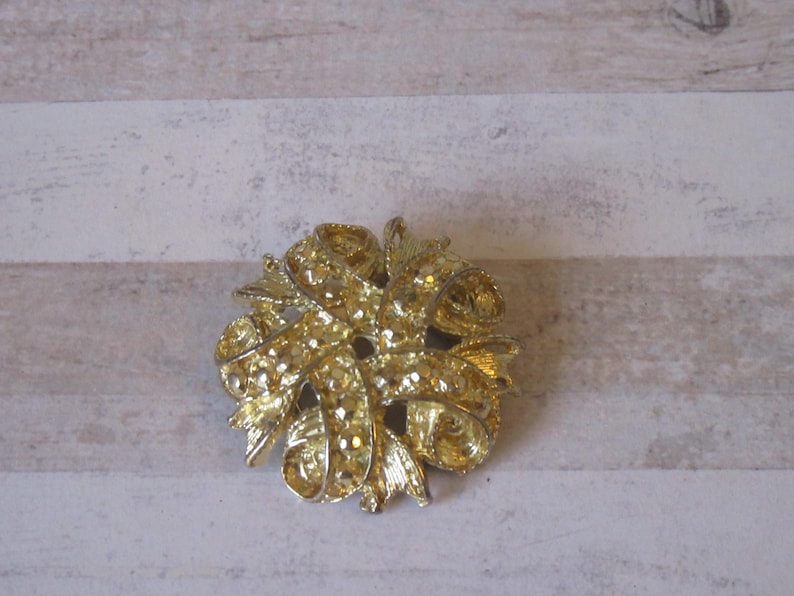 K&ouml;nnte beinhalten: Eine goldfarbene Brosche mit einem Design aus ineinander verschlungenen B&auml;ndern. Die Brosche ist filigran und detailliert, mit einer strukturierten Oberfl&auml;che.