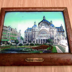 May include: A framed print of a European building with a blue dome and ornate architecture. The print is set in a dark brown wooden frame. The text "Anvers Gare centrale" is visible on the print, and "Souvenir d'Anvers" is on a small plaque.