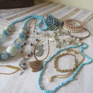Puede incluir: Una colección de varios collares, pulseras y pendientes en tonos azules, blancos y dorados. La joyería incluye collares de cuentas, una pulsera turquesa, pulseras de perlas y pulseras de cadena dorada. Los artículos están dispuestos sobre una superficie de madera clara.