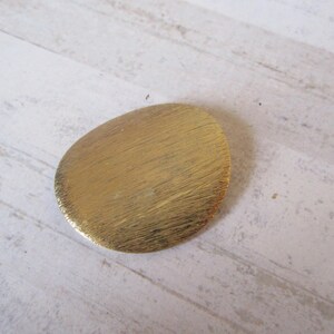 May include: A gold-colored, oval-shaped brooch with a brushed finish.