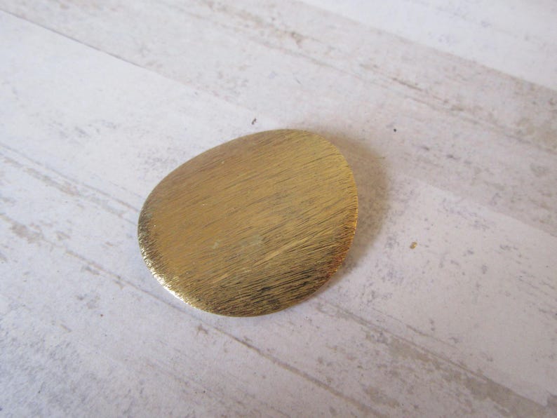 May include: A gold-colored, oval-shaped brooch with a brushed finish.