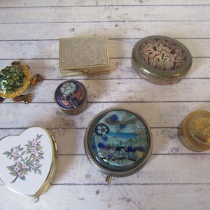 Puede incluir: Una colección de siete pequeñas cajas decorativas con varios diseños. Las cajas están hechas de metal y tienen diferentes colores y patrones. Una caja tiene forma de corazón y tiene un diseño floral. Otra caja es redonda y tiene un diseño azul y blanco. Una tercera caja es cuadrada y tiene un diseño dorado y blanco. Las cajas restantes son redondas y tienen diseños florales.