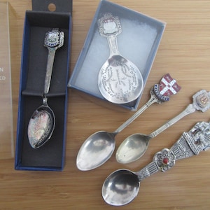 May include: A collection of four silver-plated souvenir spoons with decorative handles featuring various crests and emblems. One spoon is housed in a blue box labeled TRADITION SILVER PLATED, while another rests in a small gray box. Each spoon has a unique design.