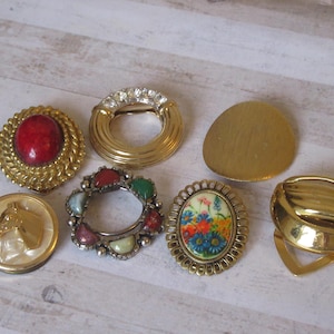 May include: Assortment of seven gold-toned brooches with various designs. One brooch features a red stone, another has a floral pattern, and another has a horse head design.
