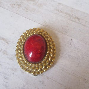 May include: A red oval cabochon gemstone set in a gold tone metal frame with a rope-like design.