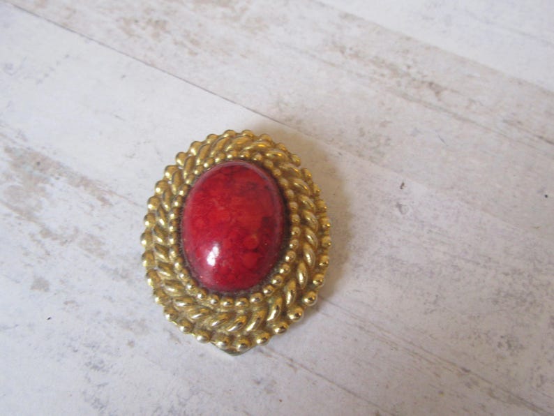 May include: A red oval cabochon gemstone set in a gold tone metal frame with a rope-like design.