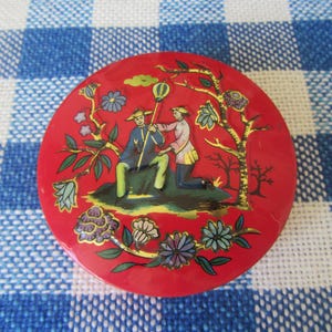 May include: A round, red decorative box with a detailed illustration. The image depicts two figures in traditional attire, surrounded by floral and tree motifs. The box has a glossy finish and is set against a blue and white checkered background.