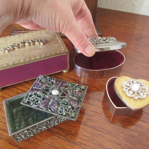 Puede incluir: Cuatro cajas decorativas con diseños intrincados. Una caja es de forma rectangular con un acabado dorado y burdeos. Otra caja es de forma de corazón con un acabado amarillo y dorado. Las dos cajas restantes son cuadradas con un acabado plateado y verde.