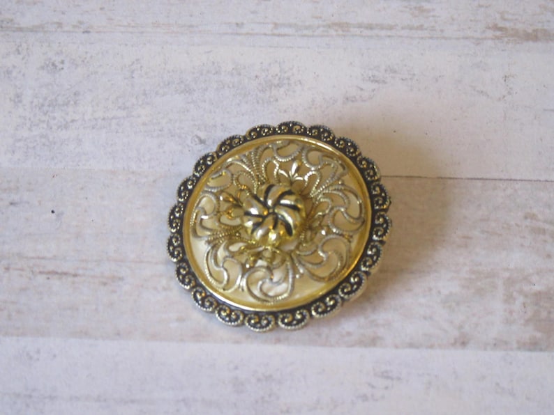 K&ouml;nnte beinhalten: Eine runde, goldene und wei&szlig;e Brosche mit einem filigranen Design. Die Brosche hat eine runde Form und eine erhabene Mitte.