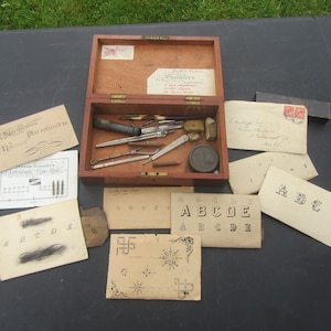 Op de afbeelding: Een open houten kist met diverse antieke gereedschappen, waaronder een klein revolverachtig instrument, een pincet en andere metalen gereedschappen. Verschillende oude papieren kaarten met gedrukte tekst en ontwerpen liggen rond de kist.