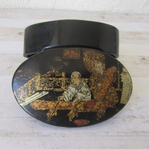 May include: A black lacquered oval box with a gold and black painted scene of two figures in traditional Japanese clothing. The box is closed and the lid is visible.
